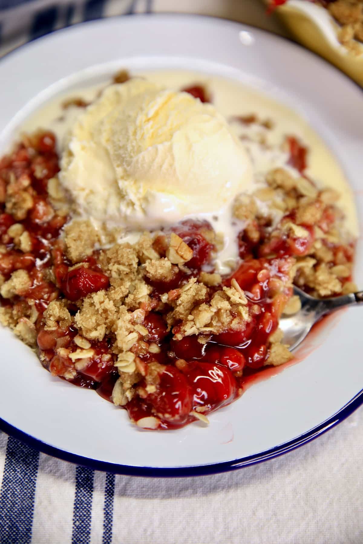 Easy Cherry Crisp is a quick and delicious semi-homemade dessert to make any day of the week that is delicious enough for company. A delicious brown sugar, oat and pecan topping over cherry pie filling and just a touch of cinnamon for a tasty fruit crisp. Cherry crisp with vanilla ice cream on a dessert plate.