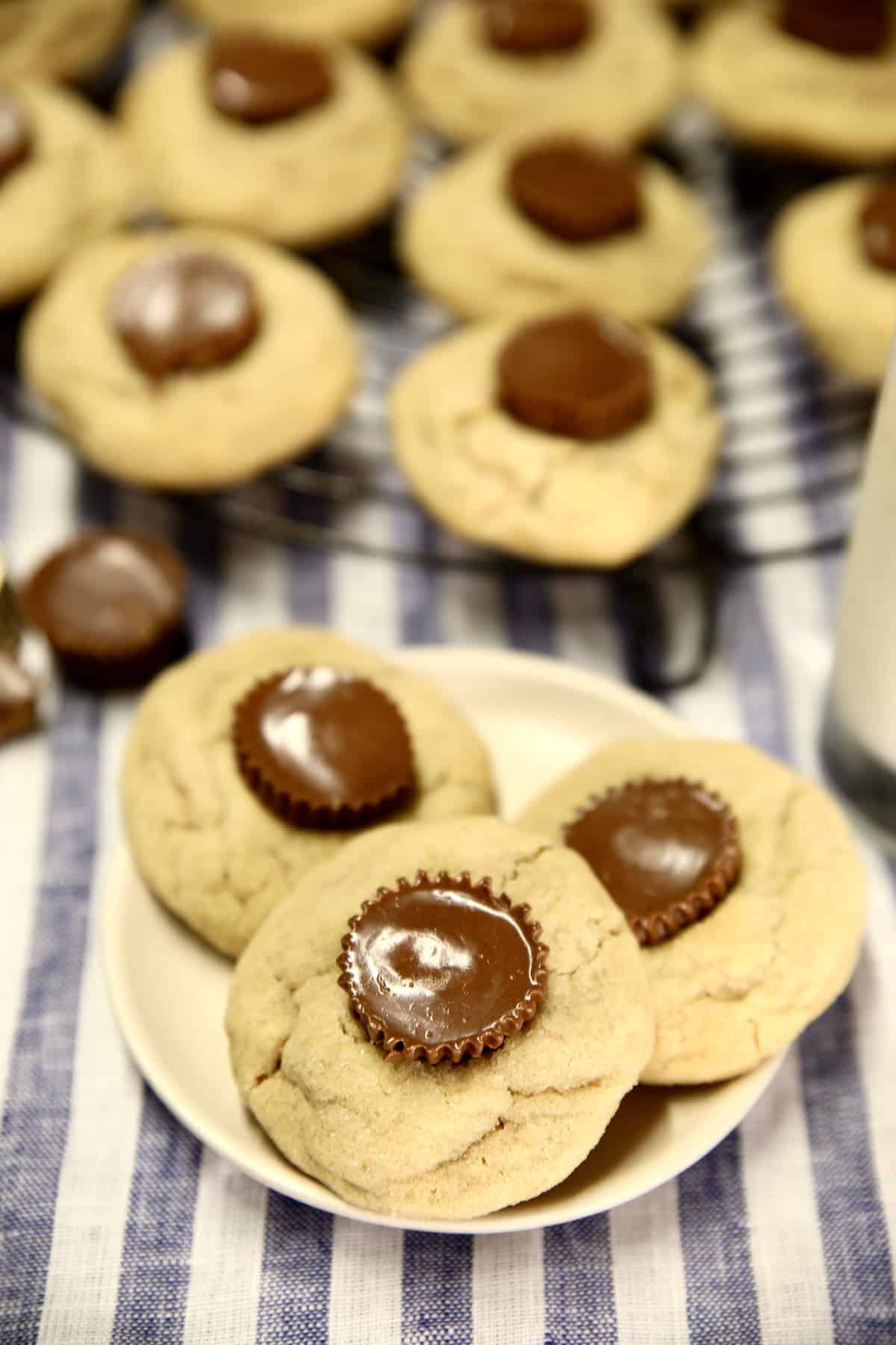3 peanut butter cup cookies on a small plate, cooking rack of cookies behind.