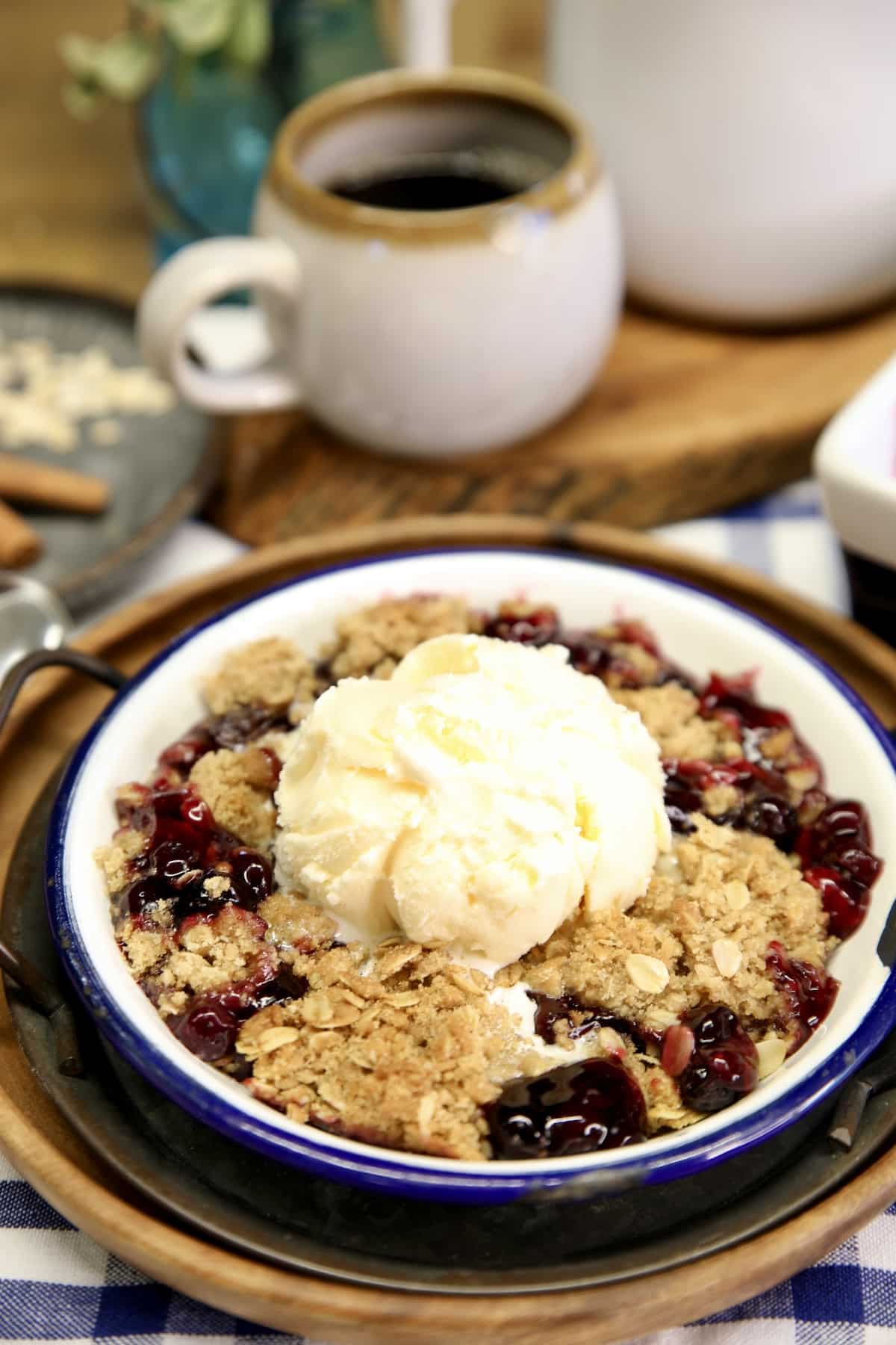 Blueberry Crisp in a bowl with vanilla ice cream. Cup of coffee in background.