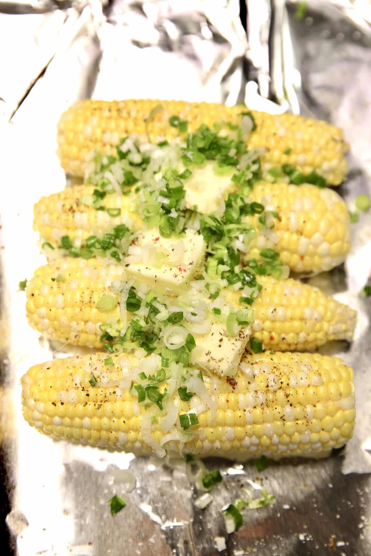 4 ears of corn on aluminum foil with butter and green onion slices.