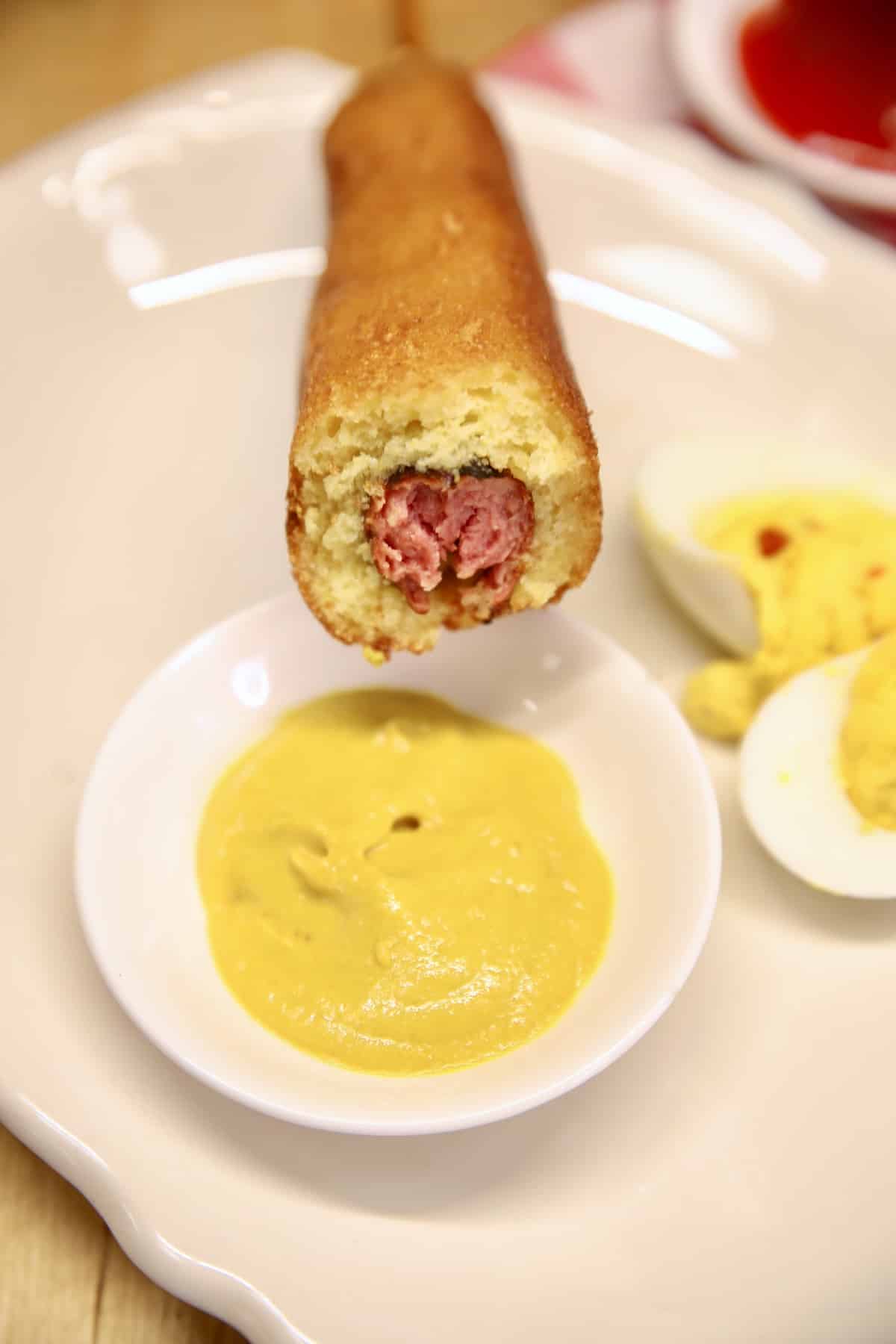 Corn dog with bite out of it, dipping into mustard.