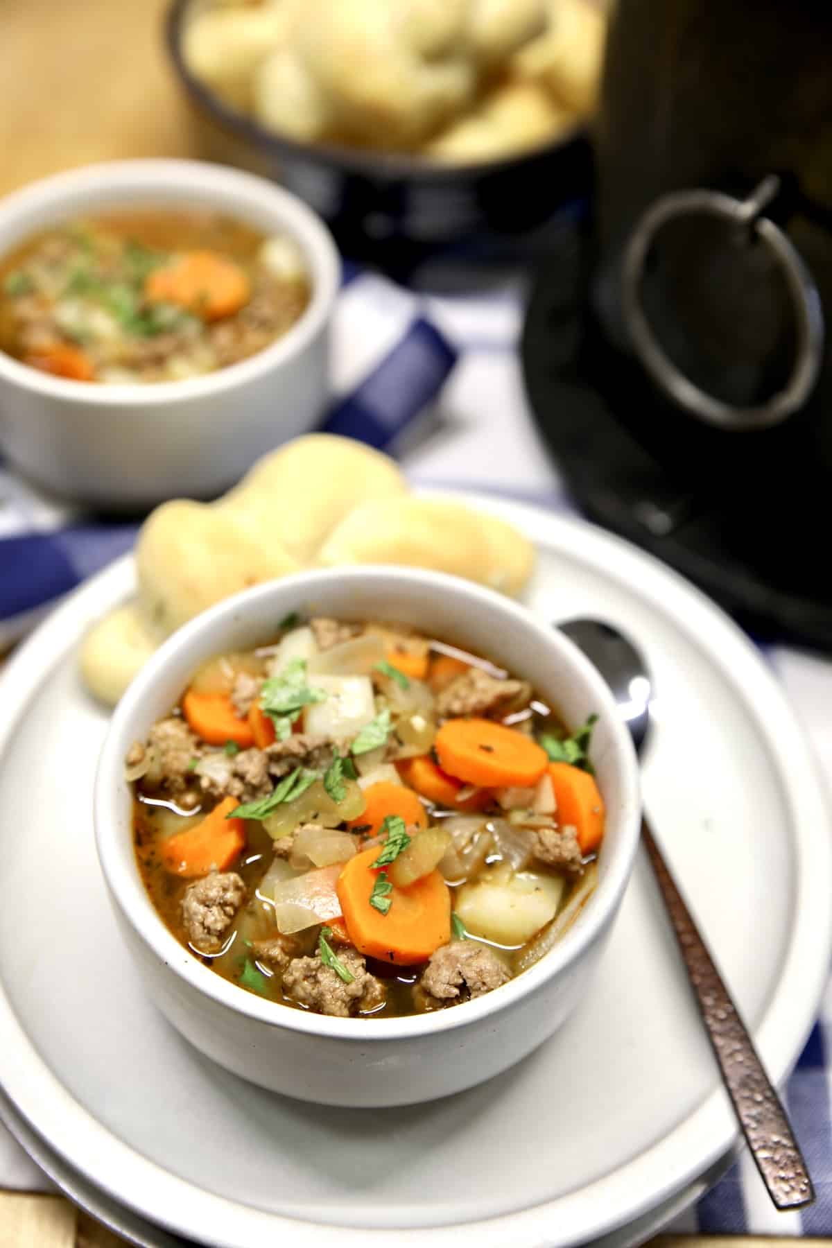 Bowl of hamburger soup on a plate with spoon and a roll.