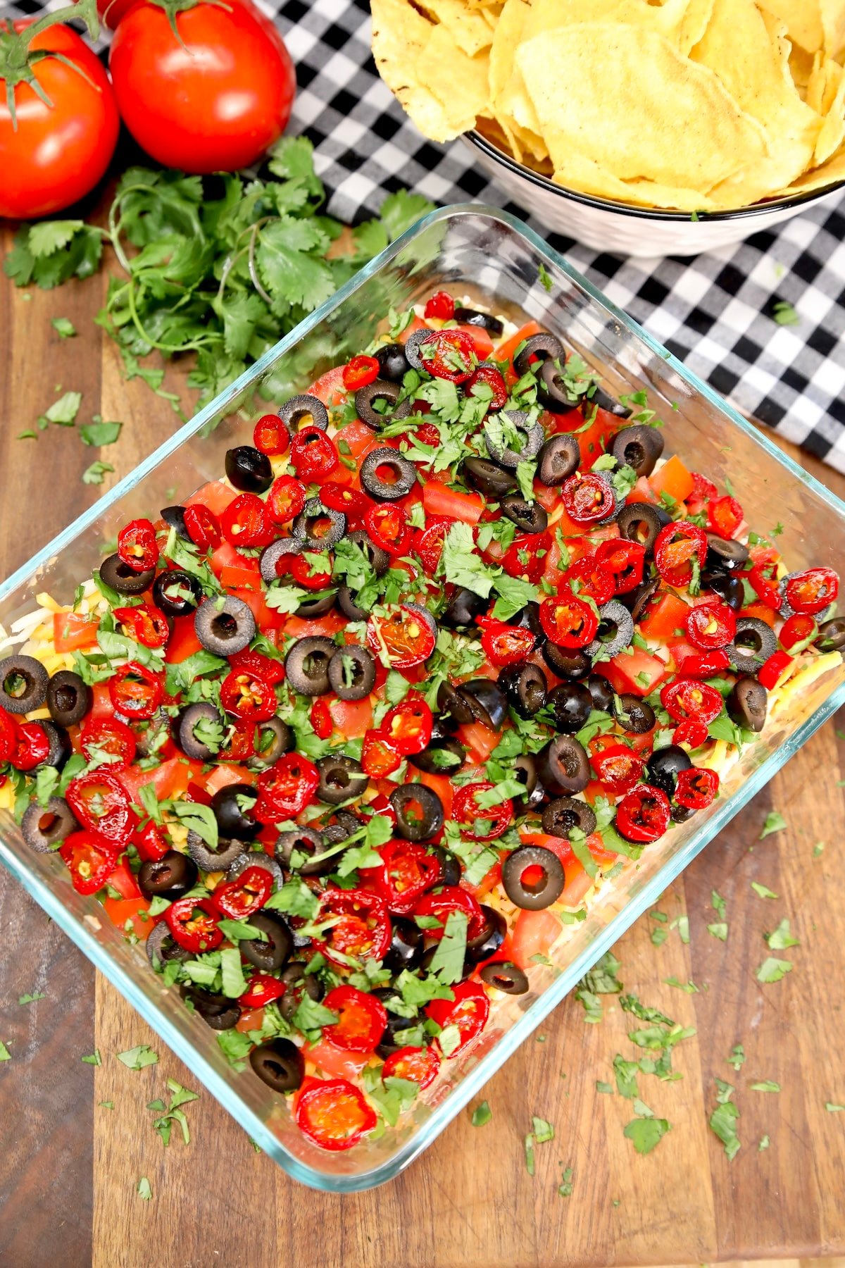 7 Layer Taco dip in a casserole dish, tomatoes, chips in background.