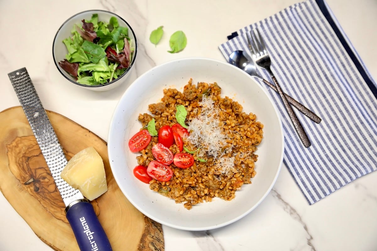 Bowl of sausage farro with tomatoes, bowl of salad, parmesan and grater.