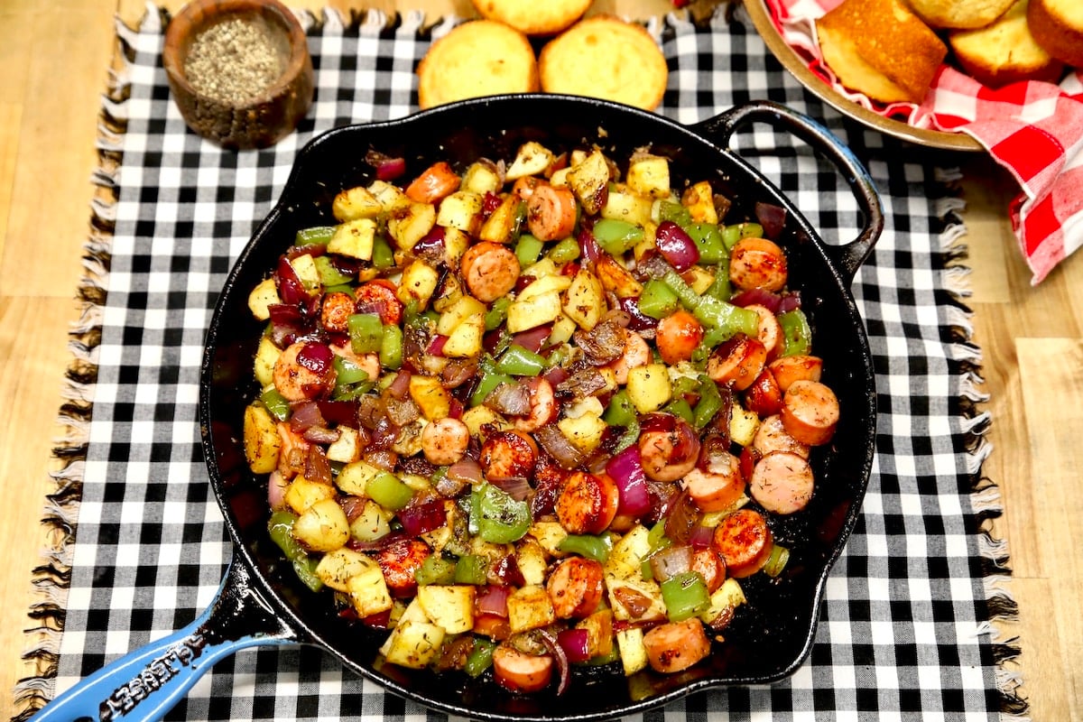 Sausage and vegetable skillet on a plaid towel, cornbread in background.