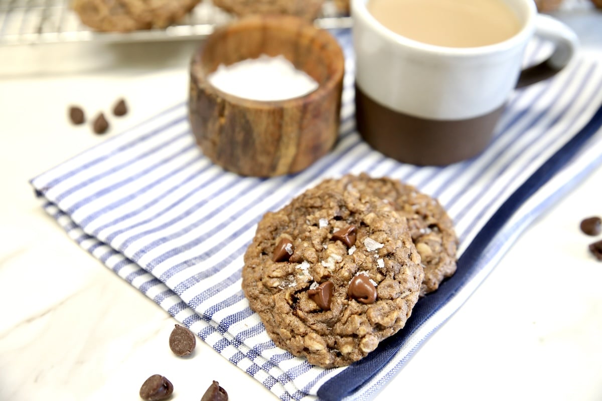 2 chocolate oat cookies with chocolate chips on stripe napkin with salt in a bowl, cup of coffee.