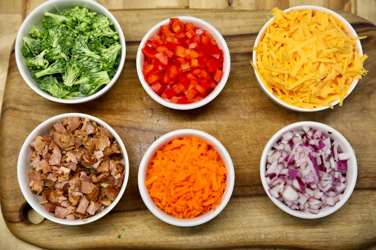 Chopped vegetables, ham, shredded cheese in bowls.