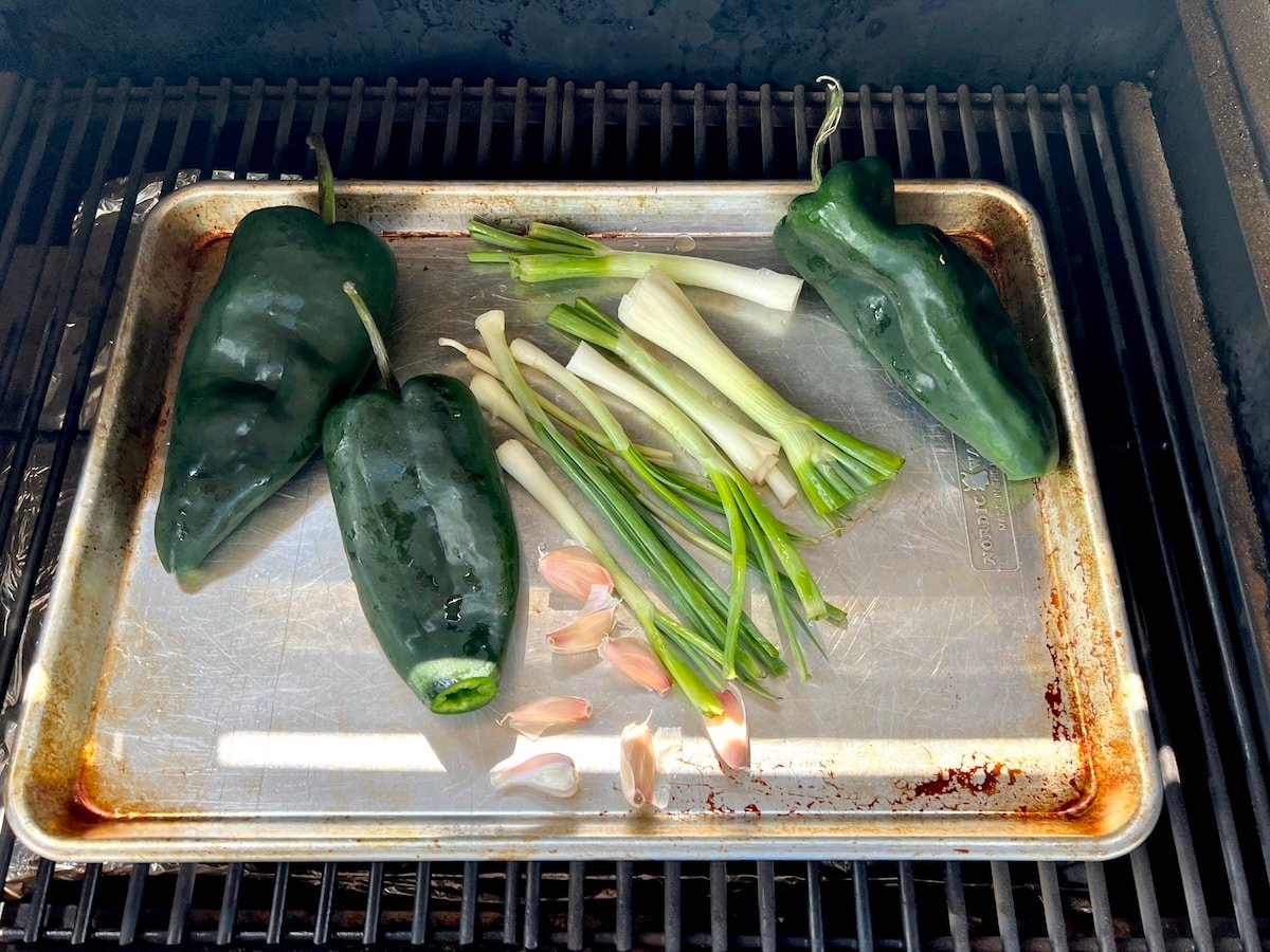 Poblano peppers, garlic, green onions on a sheet pan on a grill.