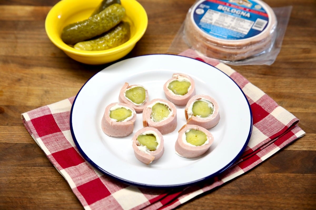 Plate of pickle roll up slices, bowl of pickles, package of bologna.